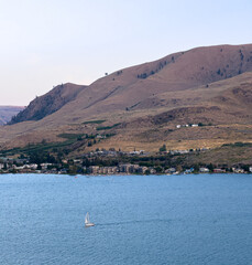 Isolated sailboat sailing across Lake Chelan in summer with Chelan houses and businesses on opposite shoreline at bottom of the beautiful hills of Eastern Washington that surround the lake.