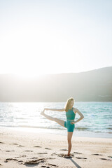 Young woman practicing standing balancing yoga poses on the beach. Ocean breeze, sunlight, and...
