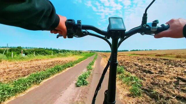 Man riding electric bicycle at high speed on dirt road in field. First person view. POV. Outdoor cycling. Blue sky with clouds