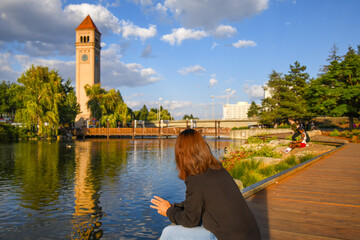 A woman sits on the riverbank boardwalk along the Spokane River at golden hour with the clock tower behind at Riverfront Park, Spokane, Washington.