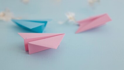 Pastel pink and blue origami airplanes on a light blue surface, surrounded by small white flowers.