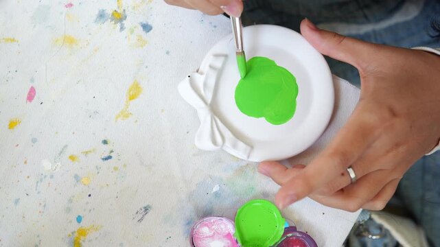 Close-up of hands painting a white ceramic ornament with bright green paint, perfect for DIY crafts, pottery decoration, and creative handmade art projects.
