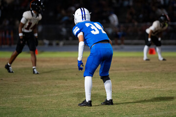 Defensive Back Football player in blue uniform number 34 crouches in ready stance under stadium lights, focused and prepared for next play during competitive night game.
