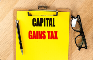 Capital gains tax text on yellow clipboard with pen and eyeglasses on wooden desk