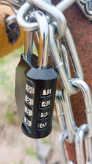 A close-up of a black combination padlock attached to a metal chain. The padlock features a numeric...
