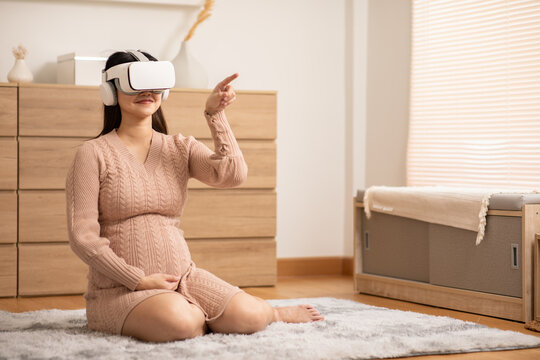 Asian Pregnant woman sitting on carpet in cozy room wearing virtual reality headset and headphones, enjoying immersive experience with relaxed and happy expression