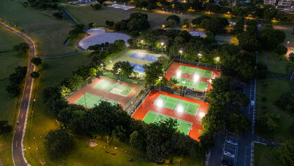 Aerial View of Illuminated Tennis Courts at Dusk Surrounded by Lush Green Trees