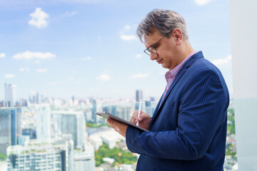 Caucasian businessman writing on tablet with stylus, standing on skyscraper, concept of real estate...