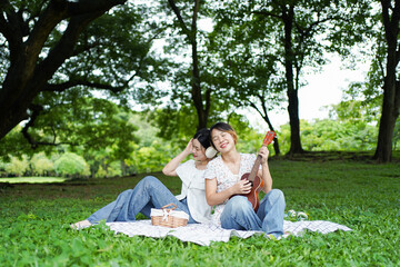 Asian siblings relaxing under a big tree in the park, playing ukulele, singing, and drinking tea...