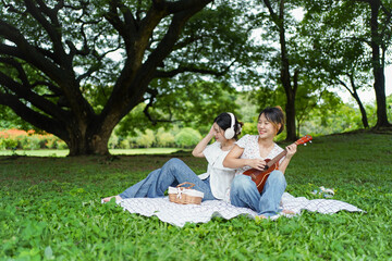 Asian siblings relaxing under a big tree in the park, playing ukulele, singing, and drinking tea...