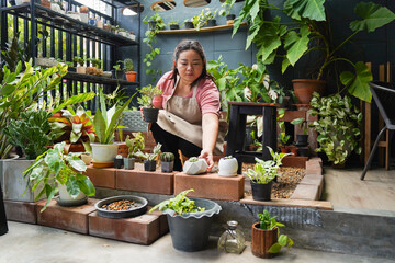 Asian woman taking care of potted plants on balcony, watering, fertilizing, and gardening with...