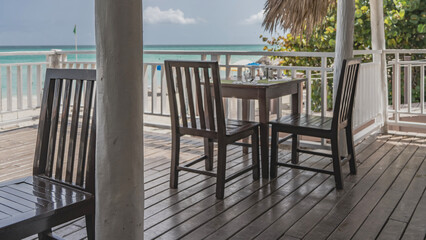 The terrace of the seaside restaurant. Tables and chairs on the wooden floor. Behind the white railings, you can see the sandy beach, turquoise ocean, green vegetation. Clouds in the sky. Cuba. 