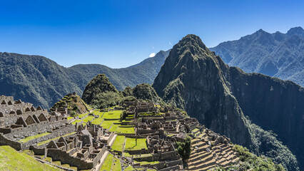 Panoramic view of the famous Machu Picchu. Ruins of a lost Inca city in the highlands. Dilapidated stone buildings, terraces, green grass. The high mountain of Wayna Pikchu against the blue sky. Peru