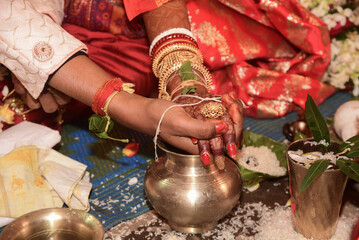 Close-up of a Hindu wedding ceremony showing the bride and groom's hands joined in a holy ritual, adorned with traditional jewelry, red attire, rice and sacred vessels.