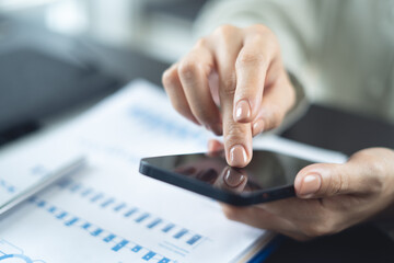 Fototapeta premium Closeup, businesswoman hand using mobile phone with business document, financial report on office table. Business woman using smartphone to calculate business data, working at office
