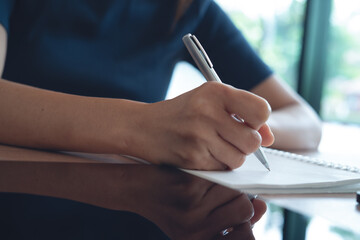 Closeup, woman hand with a pen writing on paper notebook with digital tablet on office table,...