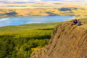 tourists sit on a high rock on the Irendyk ridge against the backdrop of the alpine lake Talkas on...