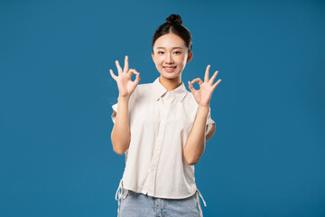 Smiling woman making 'OK' gestures with both hands against a solid blue background optimistic mood