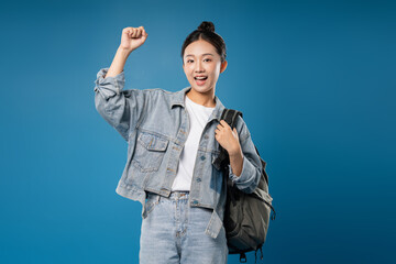 Excited student raising fist in air on a solid blue background with a denim outfit and backpack