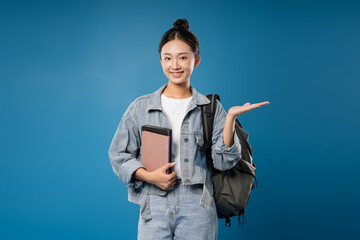 Smiling student with book backpack denim and pointing gesture on blue background School concept