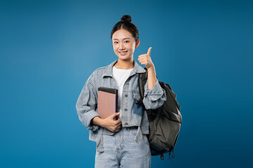 Cheerful student with backpack denim jacket book and thumbs up in front of a blue background