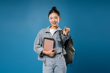 Smiling woman pointing wearing denim jacket backpack and holding books against a blue background