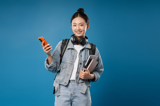 Smiling student with books and headphones using a mobile device against a blue background