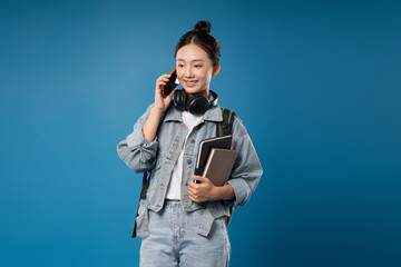 Smiling woman on phone books in hand headphones around neck against a blue backdrop