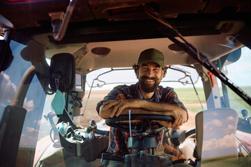 Happy agronomist driving tractor while working on a farm and looking at camera.