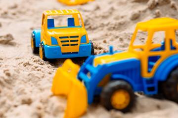 Two toy trucks are on the sand, one is yellow and the other is blue
