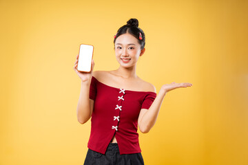 Smiling woman presenting a blank phone screen standing against a yellow background and gesturing