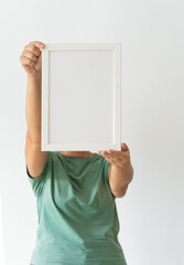 Woman in green t-shirt holding empty frame in space filled with plants against white wall. Modern...