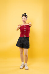 Smiling woman in red top and denim shorts pointing at the viewer against yellow backdrop