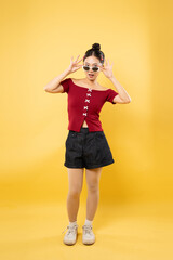 Smiling woman in sunglasses red top with white bows and dark denim shorts poses on a yellow backdrop