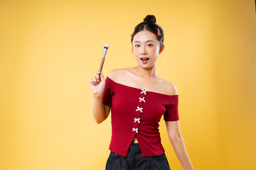 Woman with makeup brush poses against yellow background wearing red top and hair accessories