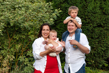 Garden portrait of cheerful parents mother holds the baby while the toddler rides on dad's...