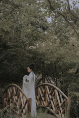 A young woman in traditional clothing stands on a wooden bridge surrounded by greenery.