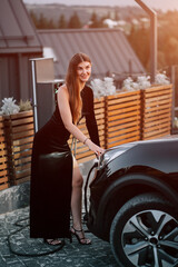 Woman dressed in stylish black outfit charging electric vehicle at sunset