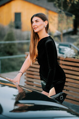 Elegant woman in a black outfit leaning on a car near a charming cabin in nature