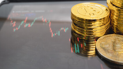 Stack of gold colored coins with Bitcoin symbol is placed on tablet displaying financial chart, symbolizing cryptocurrency trading and investment