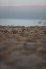 A close-up, low-angle shot of a sandy beach in soft focus, with a blurred background of the ocean and the sky at dusk.
