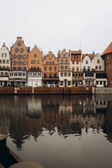 A classic panoramic view of colorful, historic buildings lining a river, with their facades beautifully reflected in the calm water under a soft, overcast sky.