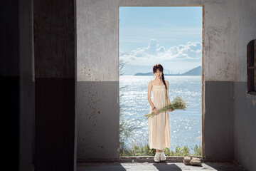 Young woman in a dress holding flowers by the water in a bright, serene setting.