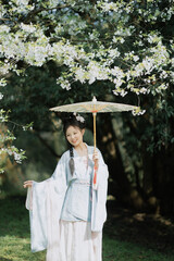 A young woman in traditional attire under flowering trees with a decorative umbrella.