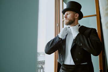 Caucasian young adult man adjusting bow tie while standing near window wearing formal suit and top...