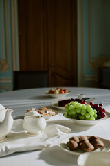 Closeup showing elegant table setting with porcelain teapot, sugar bowl, assorted cookies, green grapes, red grapes, apples on plates, background featuring wooden door