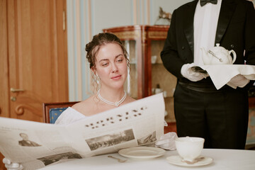 Caucasian young adult woman reading newspaper at table while Black man waiter in tuxedo serving tea on tray, elegant setting suggesting formal dining experience