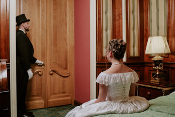 Caucasian young woman in formal dress sitting on bed facing away, while Caucasian middle aged man...