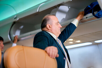 Senior businessman keep his backpack into overhead storage of airplane for business travel to...