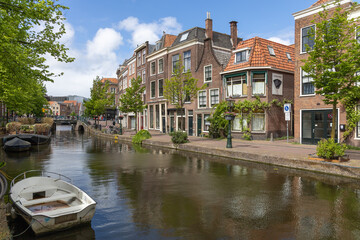 Fototapeta premium A tranquil Dutch canal scene in Leiden featuring traditional houses, green trees, and a moored boat, leading to a distant bridge under a blue sky.